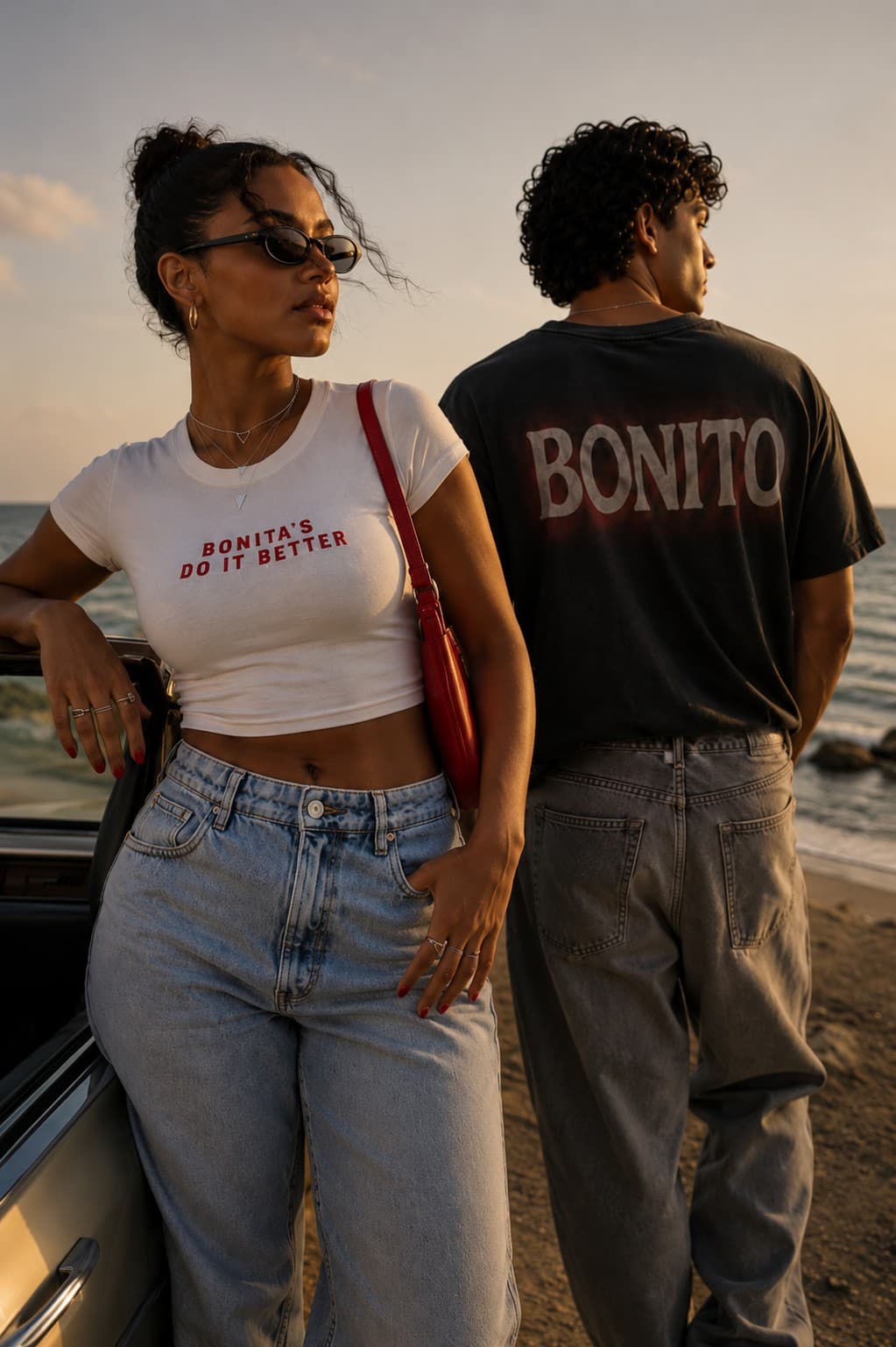Two models at the coast at sunset wearing SOUTELO: a white tee printed with Bonita's Do It Better and a washed black tee printed with Bonito